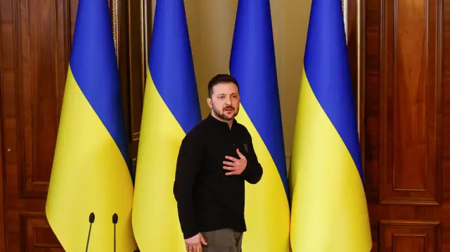 Zelensky stands with one hand on his chest with four Ukrainian flags in the background