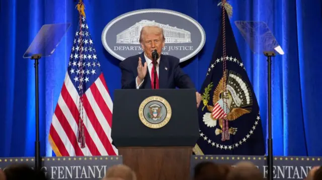 Donald Trump talking on a stage against a backdrop of US flags