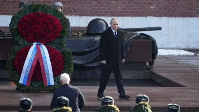 Putin standing in front of a giant wreath on a raised platform