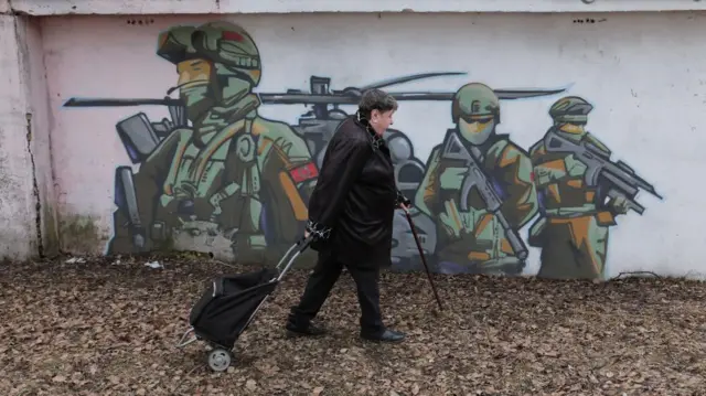 A woman wearing black and dragging a trolley walks past graffiti showing three Russian soldiers.