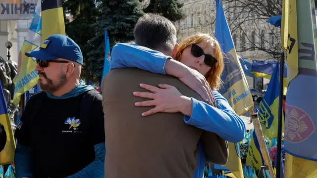 A woman wearing sunglasses hugs a man during the the commemoration ceremony in Kyiv, Ukraine. Various flags in support of Ukraine can be seen in the background