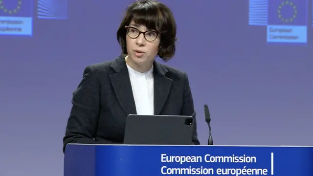 Anitta Hipper European Commission spokesperson behind a blue podium with microphone. Wearing glasses with dark, mid-length hair and a dark blazer with white shirt underneath.