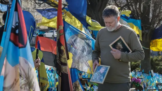 A US volunteer and advocate holds and looks down at a photo of a late US volunteer during the commemoration ceremony. He is surrounded with flags supporting Ukraine.