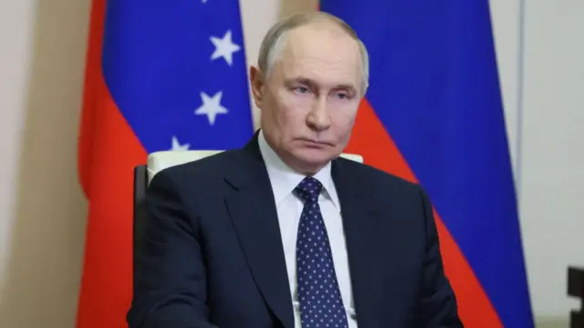 Vladimir Putin wears a suit and sits on a chair.