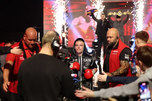 Andrew Cain makes his ringwalk dressing in black and flanked by security men