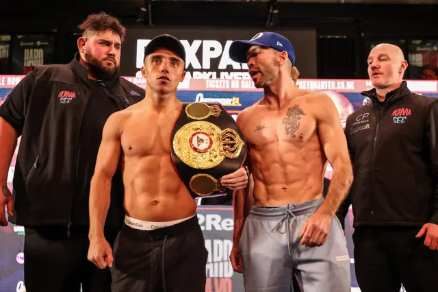 Nick Ball holds the WBA featherweight title over his shoulder and stands next to TJ Doheny on a stage at a weigh-in