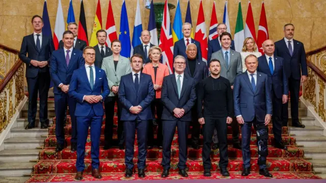 World leaders take a family photo, wearing formalwear. They arte standing on a flight of stairs in front of flags of their countries.