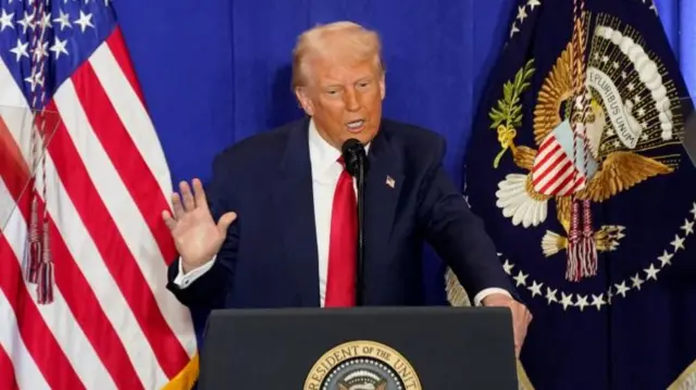US President Donald Trump in suit with red tie. Speaking behind a podium, one arm raised. US flag in the background.