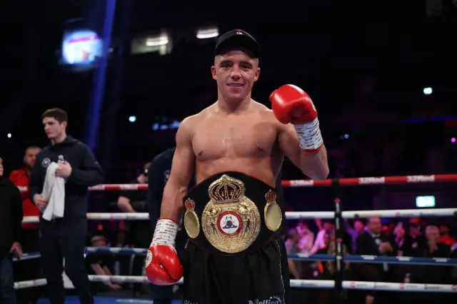 Nick Ball holds up his boxing glove and is wearing the WBA featherweight title around his waist in a boxing ring after beating TJ Doheny