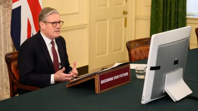 UK Prime Minister Keir Starmer sitting in front of a large monitor during a virtual call with other leaders. On the desk is a small stand with papers and a sign saying "Securing our future"