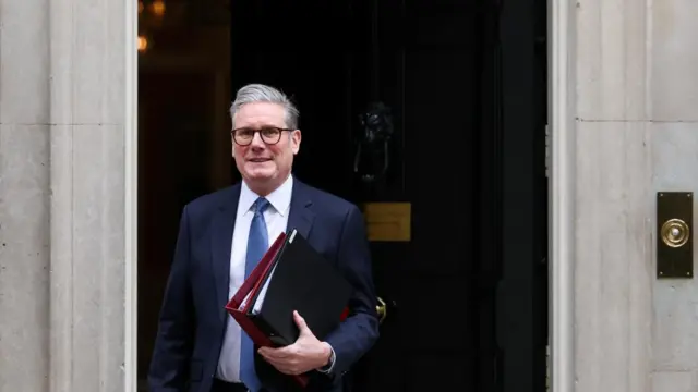Keir Starmer leaves No. 10 Downing Street holding folders and wearing a suit.