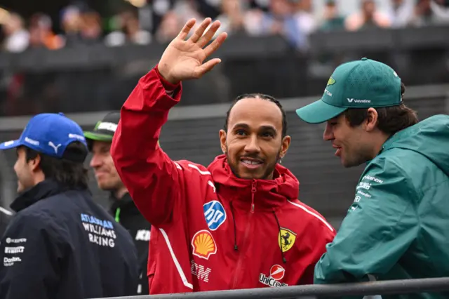 Lewis Hamilton waves to the fans in Melbourne while wearing a red tracksuit top while Lance Stroll talks to him