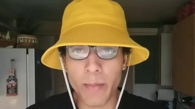 Nicholas Prosper wearing a yellow bucket hat and black glasses in a kitchen. He is looking at the camera without expression.