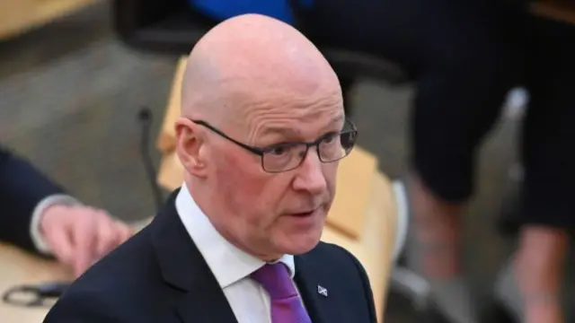 John Swinney talking in Scottish Parliament. He is facing the right, and is wearing a black blazer, white shirt and purple tie. On his left lapel, is a Scottish flag pin.