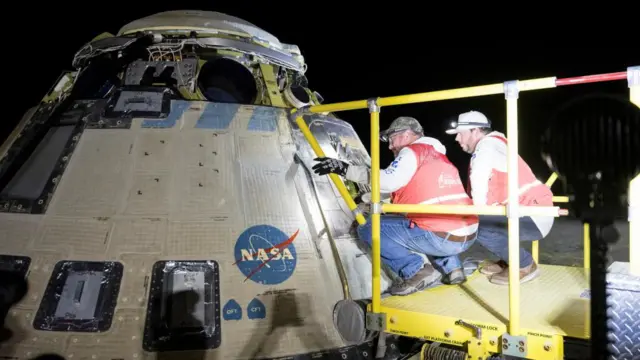 Two male engineers, who each wear blue jeans, white hoodies and orange hi-vis vests, crouch on a yellow platform inspecting the capsule, which is off-white, shaped in a cone and features the Nasa logo and many hatches and dark, rectangular sections