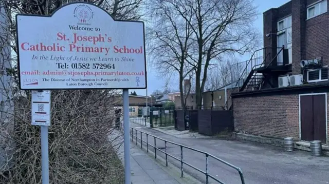 The outside of St Joseph's Catholic Primary School, showing a sign, a walkway, a brick building to the right and a school in the distance. There are metal railings in the middle.