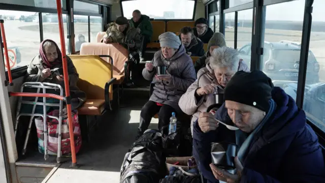 Evacuees on a bus, wearing jackets, hats and scarves, eating food.