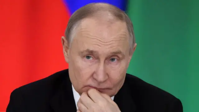 Putin headshot. He looks pensive, and holds a hand to his chin. He sits in front of a green and red background and wears a suit