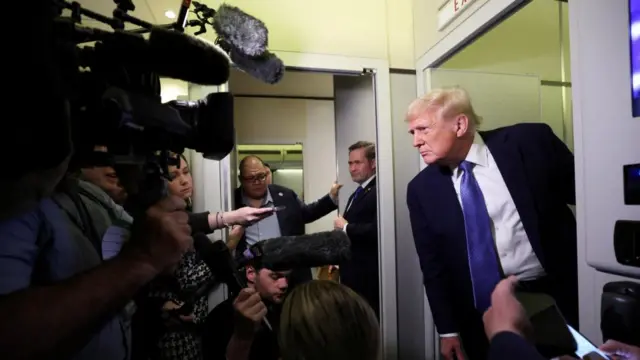 Trump stands outside of a door on Air Force One wearing a suit, surrounded by several reporters.