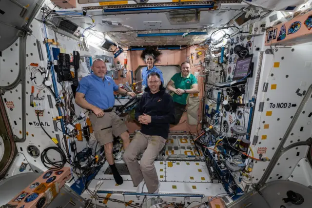 The four astronauts smile at the camera, suspended weightless in a room in the ISS with white walls covered in wires and electronics