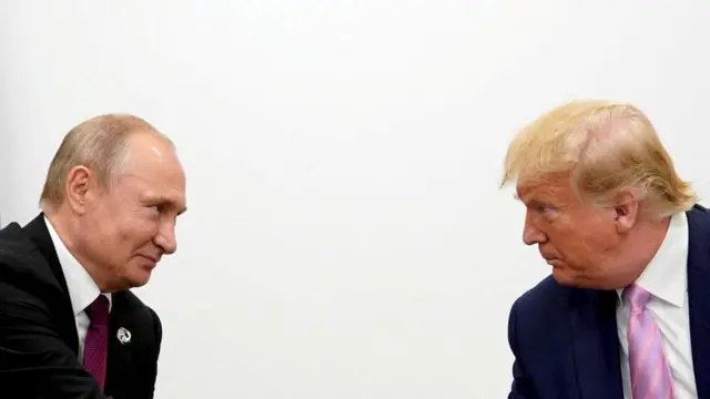 Putin, on the left, wears a black suit and has a small smile on his face, as he looks at Trump, on the right, who is wearing a blue suit with a pink tie. Trump has a more stern look on his face