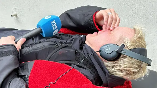 Jamie Laing holding his brow in pain whilst he lies down on a physio table holding a microphone
