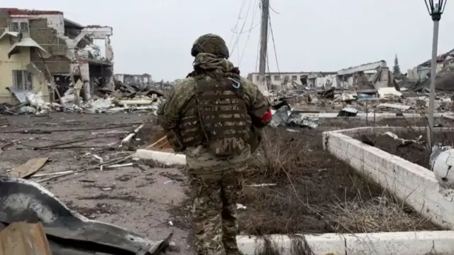 A Russian soldier, identified with red tape on his arm, walks through destroyed buildings in Loknya