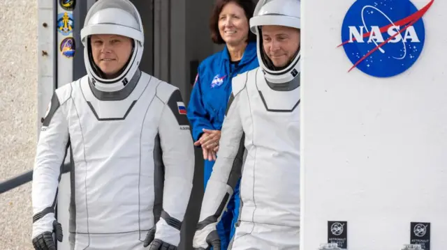 Hague and Gorbunov walk past a white wall bearing the Nasa logo wearing slim white space suits with grey detailing and matching helmets. A woman in a blue Nasa boiler suit stands behind them, seen in between them