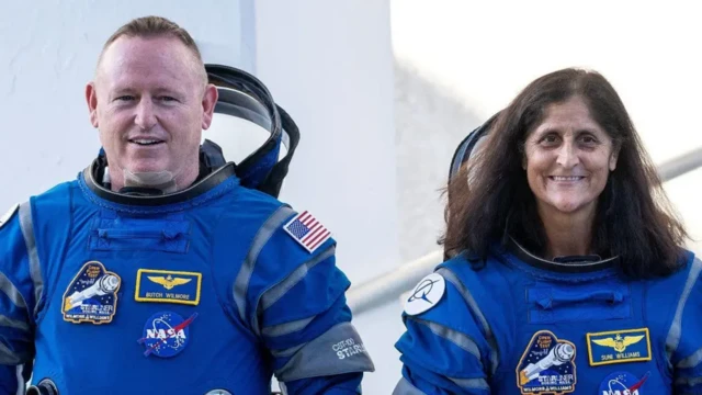 Left to right: Butch, a man with short, light coloured hair and Suni, a woman with shoulder-length brown hair, smile wearing their blue space suits