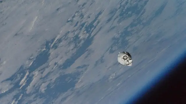 A small, round, white capsule is photographed from space above the Earth