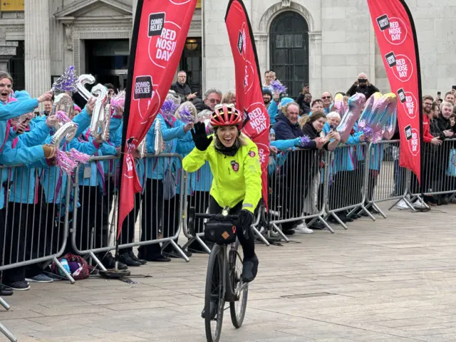 Mollie King on a bike smiling and waving with a crowd behind her cheering