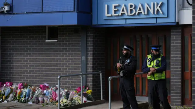 Two male police officers guarding the entrance to the Leabank block of flats. Floral tributes have been left to one side of the door.