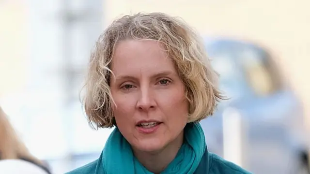 Emma Reynolds wearing an turquoise scarf around her neck and matching jacket, both of which cropped out of the image and just visible from shoulders up. She is walking outdoors.