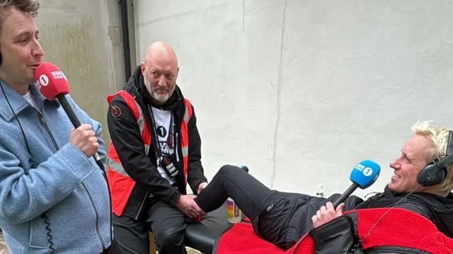 Jamie Laing receives a massage on his leg whilst speaking to Matt, they're both holding Radio 1 mics.