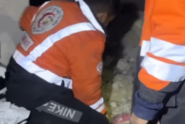 Rescue workers, wearing orange jackets, kneel over an object that cannot be identified.