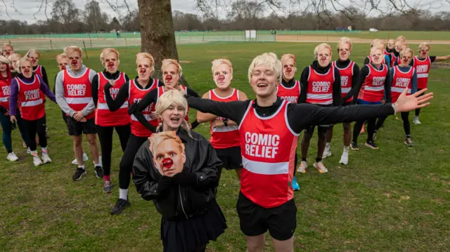 Sam and Danni with blonde wigs, in front of a crowd of people in Comic Relief tops and Jamie Laing masks