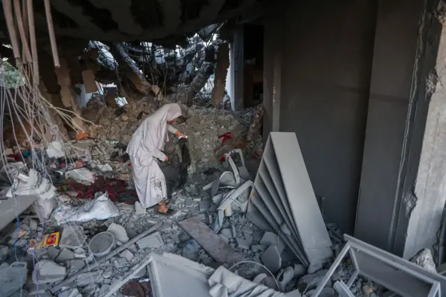 A woman in a long grey burqa looks through rubble of a building.