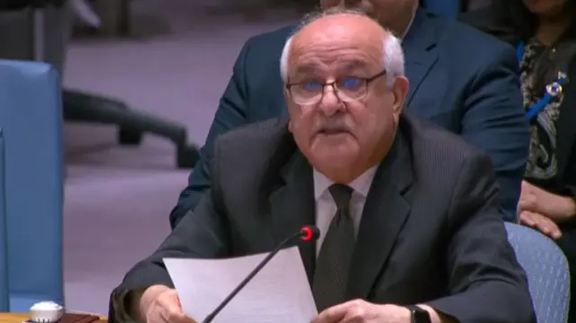 Mansour, a man with glasses, looks up as he sits at a desk in the UN behind the State of Palestine plaque, holding his speech notes on A4 paper in both hands.