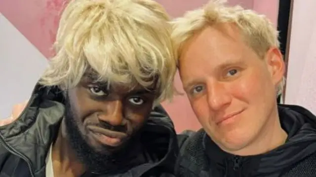 Jeremiah in a blonde wig poses for a photo with Jamie Laing