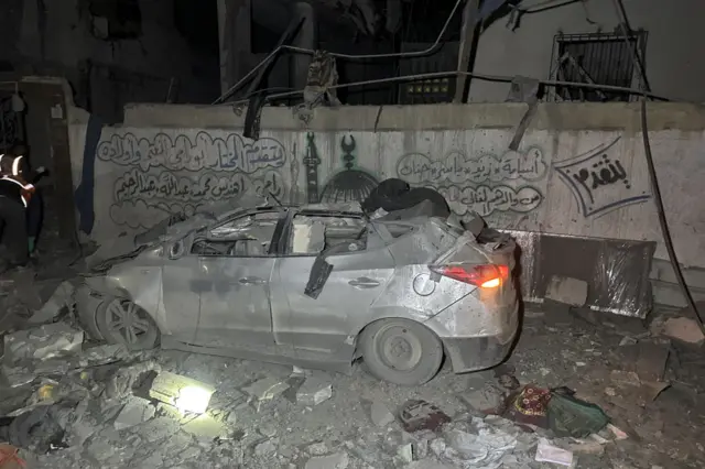 In the dark, a car sits completely destroyed. It is grey and covered in dust. There is rubbly around the car and a destroyed wall behind it