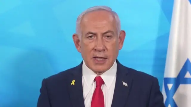 Netanyahu wears a navy suit and red tie as he speaks. He stands in front of an Israeli flag set against a blue background