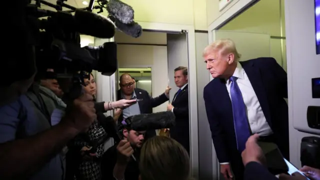 Trump speaks to reporters on Air Force One. He wears a suit and speaks in front of a group of reporters and filmmakers.