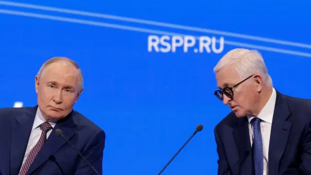Putin speaks behind a microphone alongside the Head of the Russian Union of Industrialists and Entrepreneurs Alexander Shokhin. Both are wearing suits.