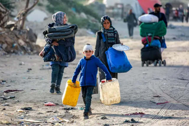 A boy walks, with a bandage around his head, holding two large water cans. Behind him are two women laden with bags and belongings, and more in the background. They walk along a dusty, dirty road, and look unhappy