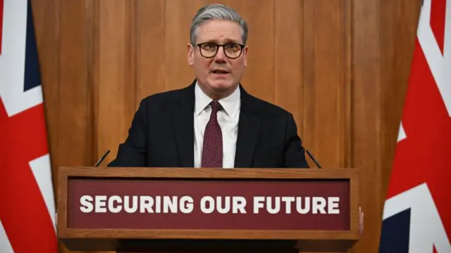 Keir Starmer stands behind a podium with microphones and flags. The sign on the podium reads: 'securing our future'.