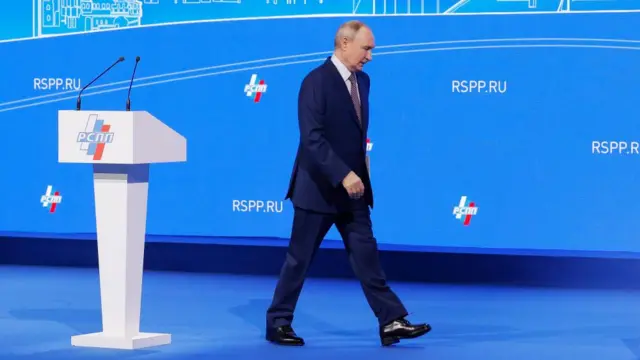 Putin walks on stage in a suit. The podium has microphones and there is a blue backdrop on stage.