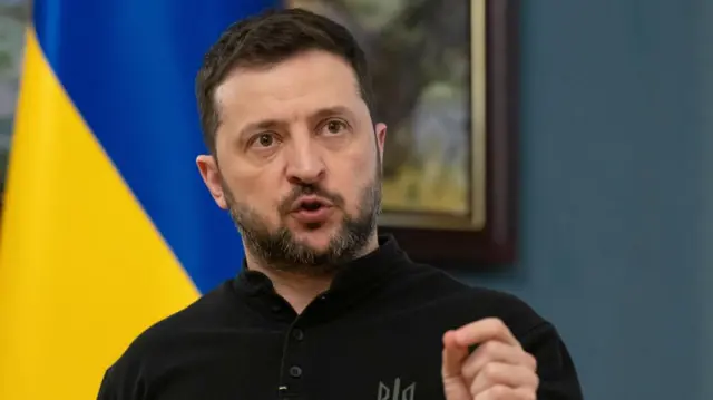 Zelensky gestures with pinched fingers as he speaks in a blue-walled room, a Ukrainian flag on his left and a painting of a landscape behind him