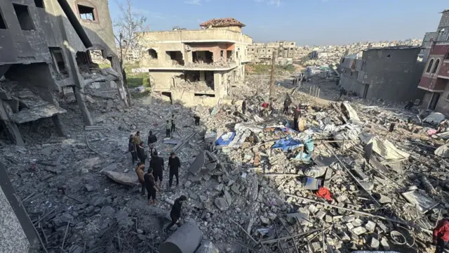 A neighbourhood flattened by strikes. Multiple buildings surround an area of total rubble, which several men stand around in. The buildings that remain standing have walls knocked out of them.