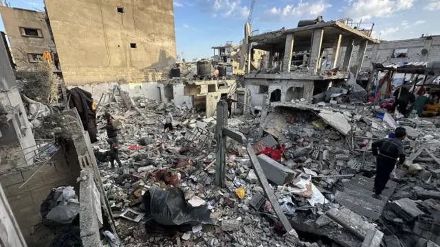 A scene of total rubble, assuming a building had been there before as you can see pillars. A  half ruined building with no walls is in the background. A man walks on steps which have no purpose now and lie on top of the mess