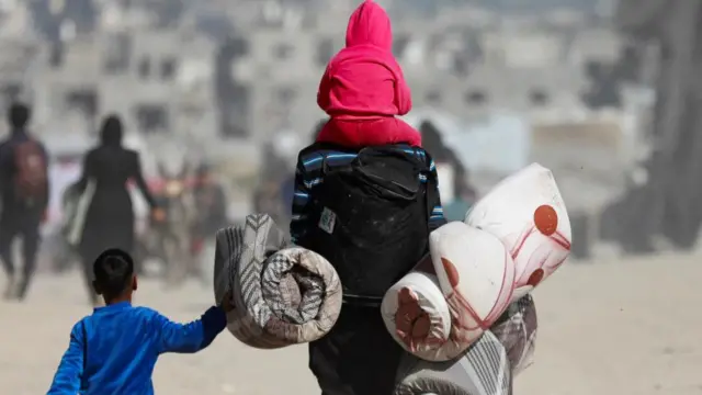 An adult carrying a child on their back and holding the hand of another as they walk with their belongings